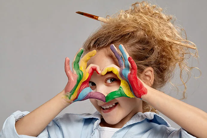 Kind formt mit bunt bemalten Händen ein Herz vor dem Gesicht