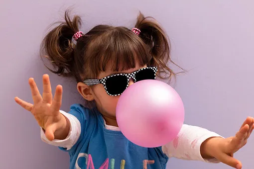 Kind mit aufgemalter Sonnenbrille pustet eine rosa Kaugummiblase